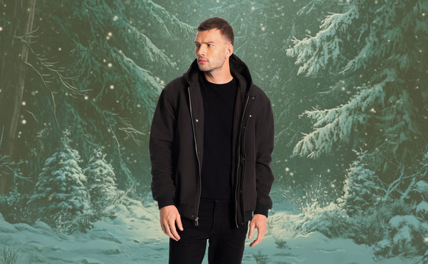 Man wearing a black Hestiia bomber jacket standing in front of a snowy forest background