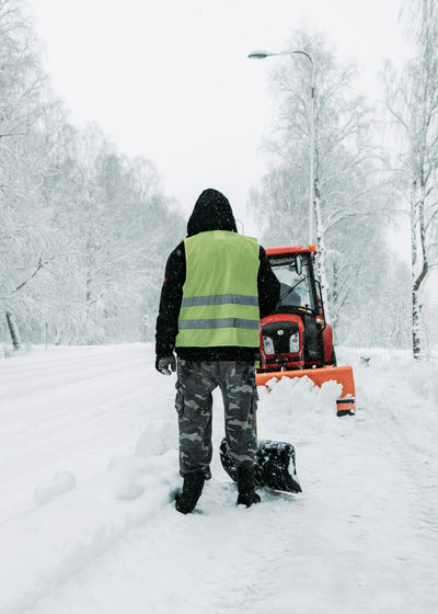 Person in a high-visibility vest and a black jacket with hood up holding s hovel in front of a snow plow in a snowy landscape