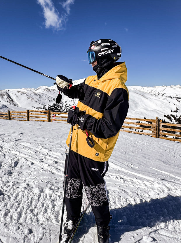 Person in a yellow and black Hestiia 7k3 jacket with a helmet and goggles, standing on a snowy slope.