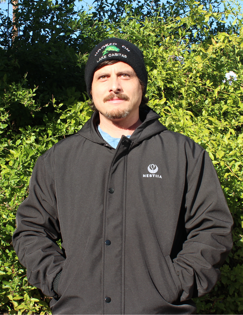 A man wearing a black Hestiia  jacketand a black beanie stands outdoors in front of green bushes. The jacket has a Hestiia logo on the left chest.